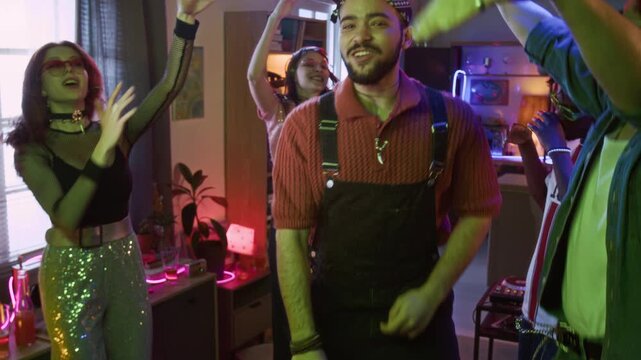 Medium portrait shot of cheerful young adult biracial man hyping up crowd dancing to music while friends raising hands up and singing along song at home party