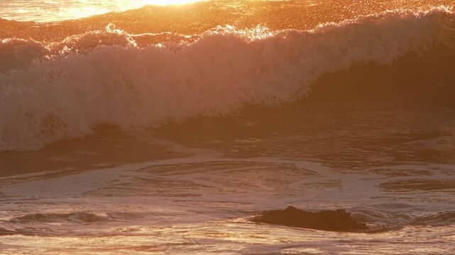 Waves crash on the shore at Costa da Caparica, Portugal. The sun backlights the water, creating a beautiful display of splashes and reflections in slow motion during sunset.