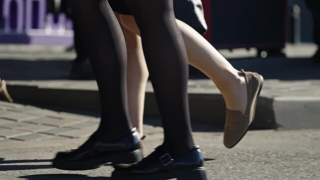 Close-up of varied footwear in motion on a sunlit urban sidewalk, showing heels, flats, and black tights with chunky loafers as pedestrians stride across pavement during a commute