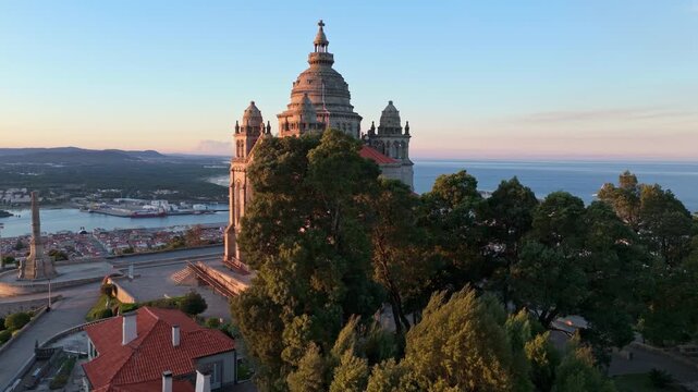 Drone reveal shot starting behind trees and moving towards the Santa Luzia Sanctuary in Viana do Castelo, Portugal at sunrise. Reveal orbit shot.