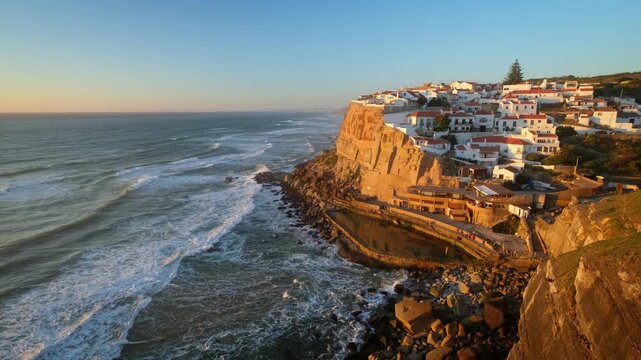 Azenhas do Mar, a fishing village in Portugal located on a cliff by the Atlantic Ocean. The sun sets over the coastline, casting warm light over the village and sea.