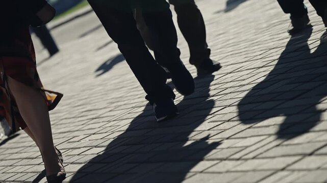 Pedestrians stride across a sunlit paved sidewalk, their long shadows stretching diagonally as a low-angle view captures motion and blur, evoking the rhythm of an urban commute.