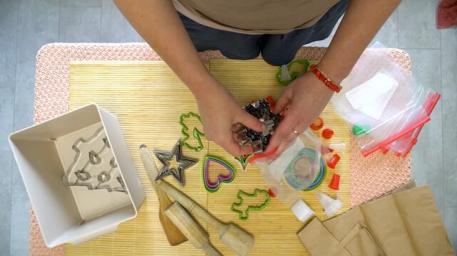 Hands are busy organizing cookie cutters and baking supplies on a wooden table. The setting is a well-lit kitchen, ideal for creative baking activities.