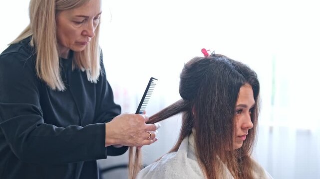 A hair stylist performs keratin hair straightening on a woman's hair using a flat iron. The process takes place in a beauty salon with careful attention to detail.