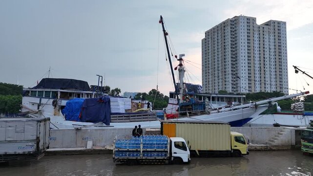 Traditional wooden cargo ships and modern trucks operate at a flooded harbor terminal during high tide, showing the busy logistics and maritime trade activities near a tall urban apartment building.