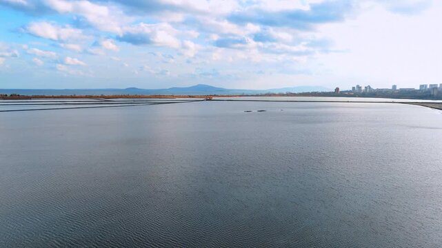 Calm salt evaporation ponds under a blue cloudy sky. Wide aerial perspective of still water in salt pans with distant mountains.