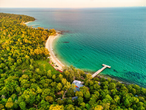 Aerial view of lush green canopy meeting the turquoise sea, with a pristine beach and a wooden pier stretching into the water, Preah Sihanouk, Sihanoukville, Cambodia.