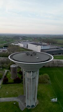 Aerial view of a concrete water tower standing tall amidst green fields and industrial buildings, contrasting with the vast sky, Ath, Wallonia, Belgium.