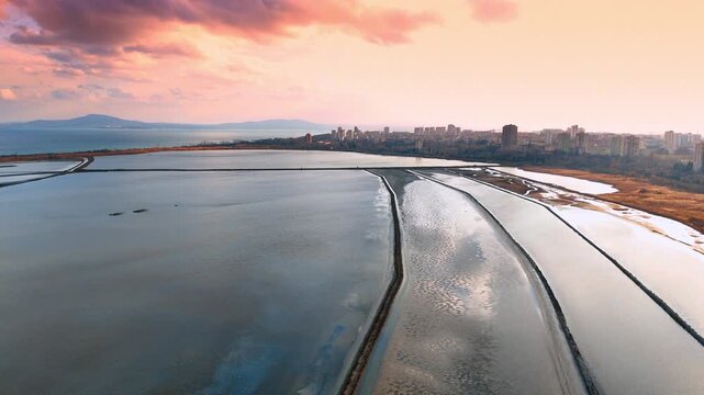 Sunset over salt ponds with city skyline in background. Aerial view of salt evaporation fields during golden hour with urban horizon.