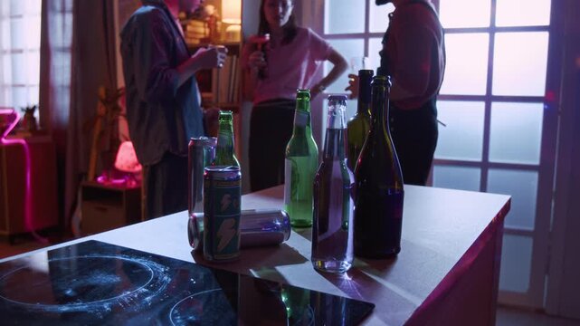 Rack focus shot of three cheerful young adults chatting in kitchen holding glasses of beverages while empty bottles resting on counter during home party