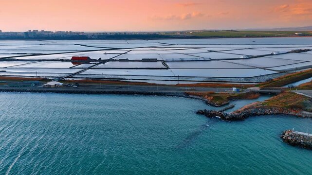 Wide aerial shot of salt pans and sea harbor entrance. Industrial salt ponds separated by dams with a small pier in foreground.
