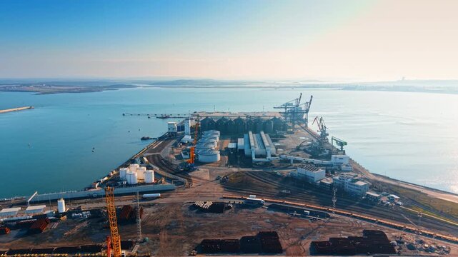 Aerial view of industrial port terminal with bulk storage silos. Wide drone shot of sea port infrastructure, grain silos, fuel tanks, and cargo berths under clear sky.