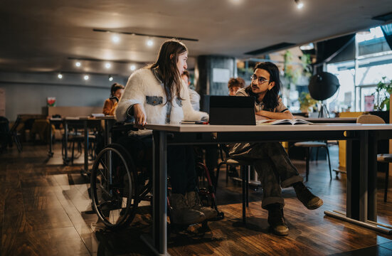 A young wheelchair user and a colleague work together at a table with a laptop. The pair converse and study in a relaxed, accessible coworking environment.
