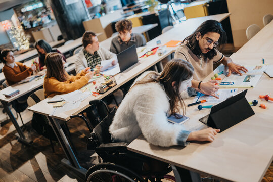 A diverse group of college students studying and collaborating at long tables in a classroom. Peers work together with laptops, books and notes in a shared study session.