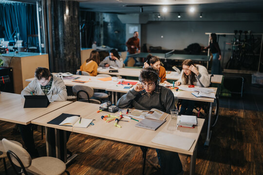 A group of students studies and works quietly at communal tables with books, laptops and stationery. Young people focus on reading, writing and digital work in a casual co-learning environment.