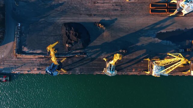 Top down aerial view of yellow port cranes and coal. Port cranes and piles of coal on the dock next to green water.
