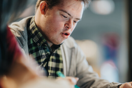 Young student studying intently while writing with a pen at a table in an indoor learning space. A focused student works on a task, showing concentration and engagement in a casual classroom