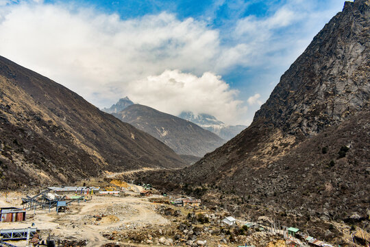 Rugged Himalayan valley with rocky terrain, scattered buildings, and distant snow peaks under dramatic cloudy sky in North Sikkim.