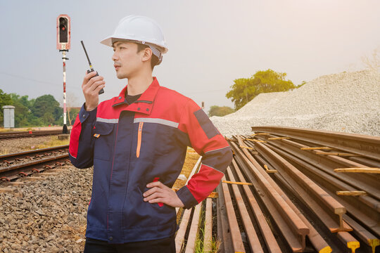 An Asian railway engineer in a safety helmet and uniform using a walkie-talkie to communicate at a train track construction site with steel rails and ballast piles.