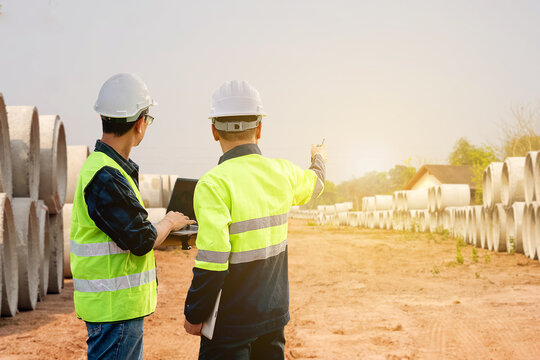 Two Asian engineers in hard hats and safety vests are collaborating on site, with one using a laptop and the other pointing, amidst large concrete drainage pipes and dirt ground.