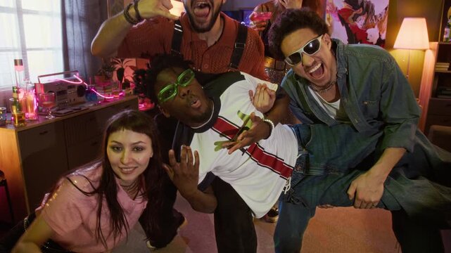 Dolly in portrait shot of multiethnic young adults with sunglasses making devil horns gestures and pulling faces while holding friend on hands and cheering while partying at home