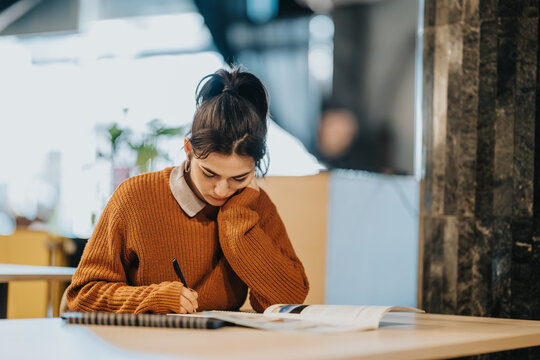A young student intently writes in a notebook while seated at a table in a cafe. The scene shows a focused student studying and taking notes in a relaxed public workspace.