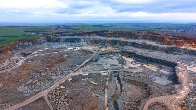 Wide aerial panorama of large scale industrial stone quarry. Panoramic view of a deep open pit mine with tiered rock walls under a cloudy sky near green fields.