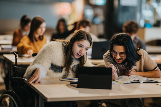A female student in a wheelchair and a male classmate study together using a tablet and open book. They collaborate over digital material and notes at a shared desk in a classroom setting.