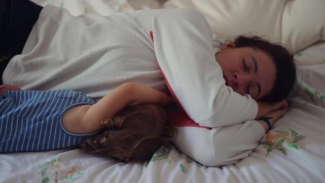 Mother sleeping on bed with arm wrapped protectively around toddler child capturing tender family closeness and quiet moment of parental fatigue in home bedroom