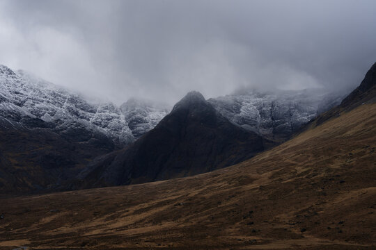 View of rugged, snow-dusted peaks shrouded in mist rise above the earthy brown slopes, creating a dramatic contrast, Isle of Skye, Scotland.