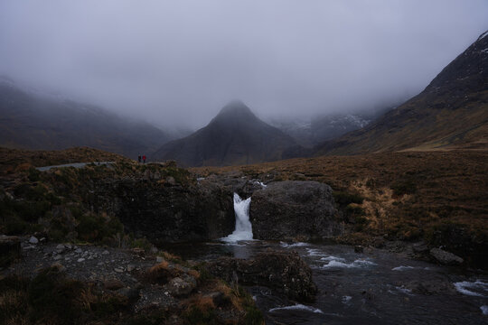 View of a cascading waterfall plunges amidst dark, brooding rocks, framed by misty Cuillin mountains under a heavy sky, Fairy Pools, Isle of Skye, Scotland.