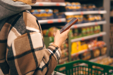 Fototapeta premium Woman shopping in a supermarket, checking product information and comparing prices on her smartphone, navigating retail aisles with a digital shopping list application