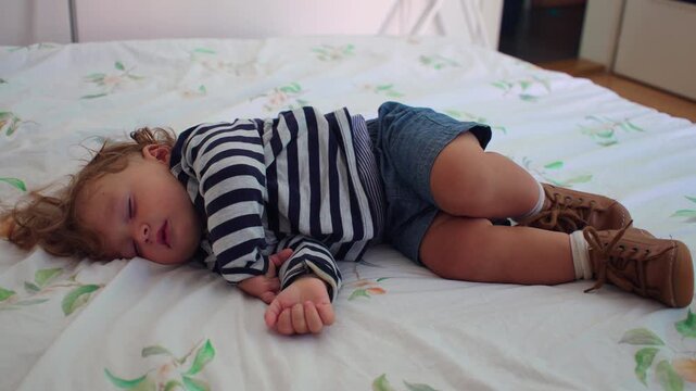 Toddler asleep on bed in curled position wearing small shoes, peaceful childhood rest moment suggesting exhaustion after play inside quiet family home bedroom