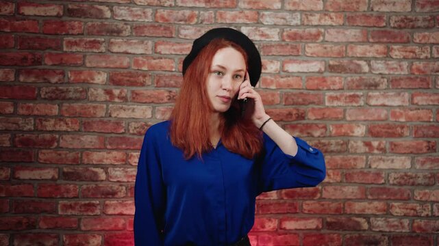 A front view of annoyed caucasian young redhead woman in a blue blouse and black beret, talking aggressively and brazenly on the phone against a brick wall. Unfriendly conversation with scammers