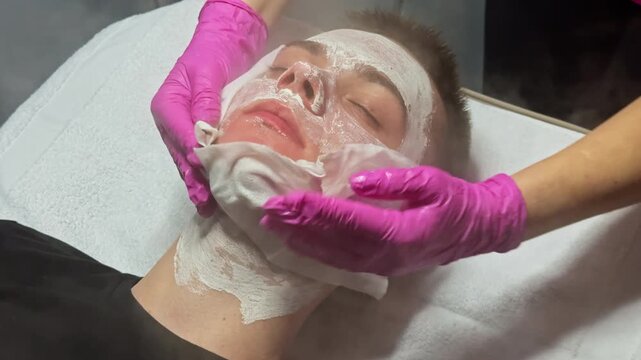 A client lies on a treatment bed in a skincare salon while a professional applies a facial mask. The client wears a simple top and has his eyes closed. Hands with gloves carefully place the mask.
