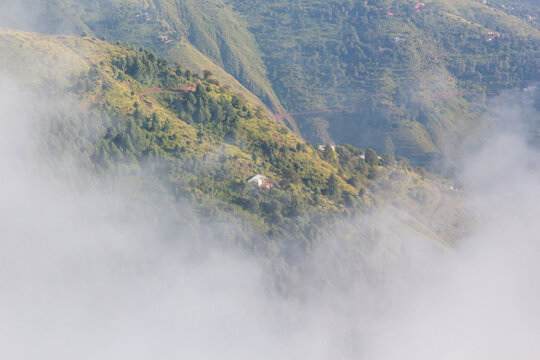 View of fog rolling across the lush, green mountainside, partially obscuring a small building nestled among the trees, Pir Chanasi, Azad Kashmir, Pakistan.