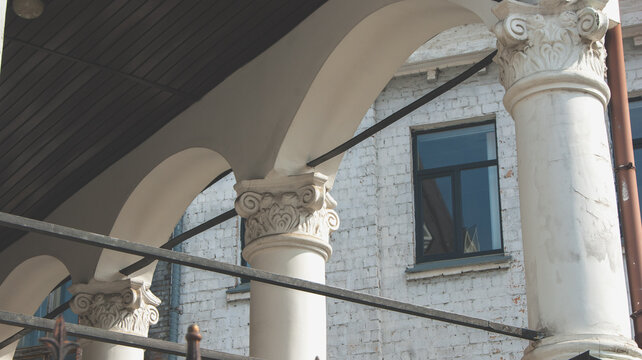 Classical architectural detail featuring white columns with Ionic capitals and arches. Historical building facade with brick walls and large windows in the background.
