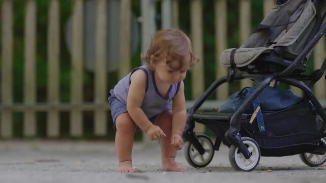Toddler trying to stand up on gravel beside stroller then losing balance and falling gently on bottom, playful childhood learning moment showing persistence curiosity and growth