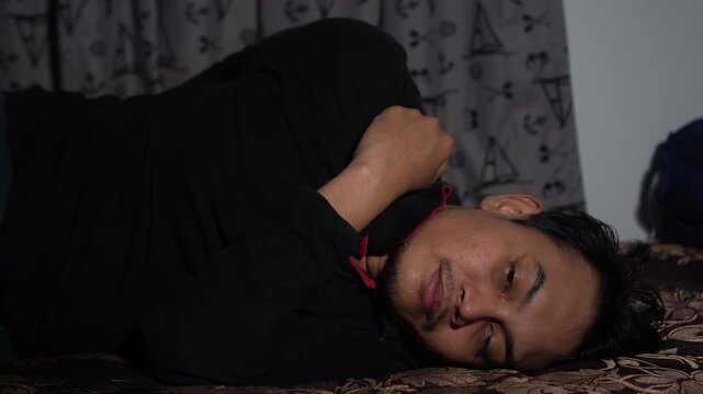 A man lying on his side in bed, waking up and immediately checking his smartphone in a dark room. Concept of digital addiction, morning routine, social media scrolling, or insomnia. Moody indoor light