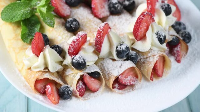Thin pancakes filled with fresh berries, topped with whipped cream, strawberries, and blueberries, dusted with powdered sugar and mint. Close-up of the dessert on a white plate. Horizontal video. Circ