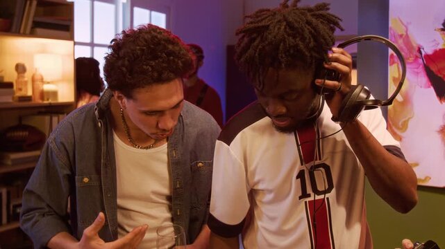 Tilt up shot of young adult African American man showing male friend how to mix tracks on DJ controller while hanging out together at home party