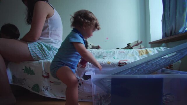 Curious toddler rummaging through toy storage box on bedroom floor while mother sits nearby on bed, playful moment of discovery, childhood curiosity, home parenting lifes