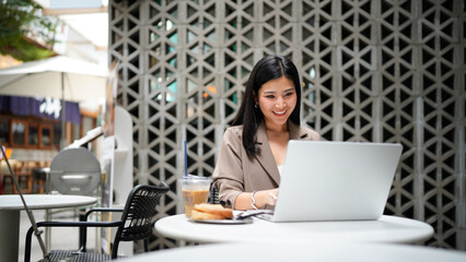 Fototapeta premium Happy young business woman worker or student working on laptop computer outdoors in outdoor cafe. Professional businesswoman employee sitting at cafe table relaxing browsing internet online