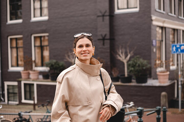 Fototapeta premium Confident and smiling young woman wearing a classic beige trench coat standing on a European city street with historic architecture, bicycles and sunglasses on her head during a cozy autumn day