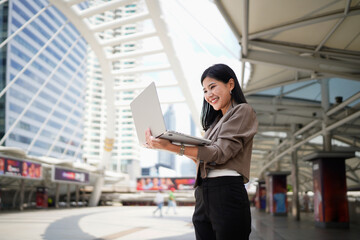 Fototapeta premium Successful Asian businesswoman smiling while working on laptop standing in downtown surrounded by modern skyscrapers in the urban financial district