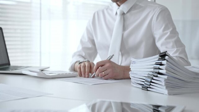 Businessman working at a desk, reviewing official documents, writing with a pen, and using a calculator, managing a stack of organized paperwork in a professional office environment. Audit and taxes