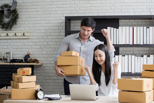 Joyful couple packing cardboard boxes in warmly lit home office, surrounded by bookshelves and festive Christmas decor, excitedly celebrating successful shipment.