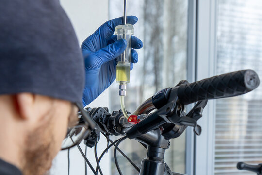 A professional mechanic wearing protective nitrile gloves performs a hydraulic disc brake bleeding procedure on a black bicycle handlebar assembly to ensure optimal stopping performance