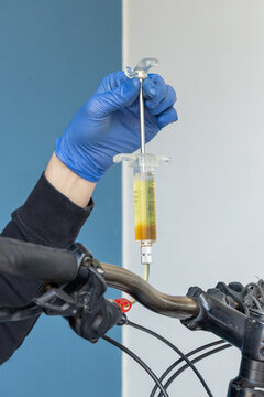A professional bicycle mechanic wearing blue protective nitrile gloves uses a specialized syringe kit to perform a hydraulic disc brake bleed on a mountain bike handlebar in a repair workshop