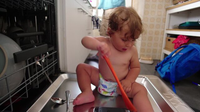 Toddler playing with kitchen utensils while seated inside dishwasher, capturing independence, experimentation, and playful misuse of household space in authentic home environment